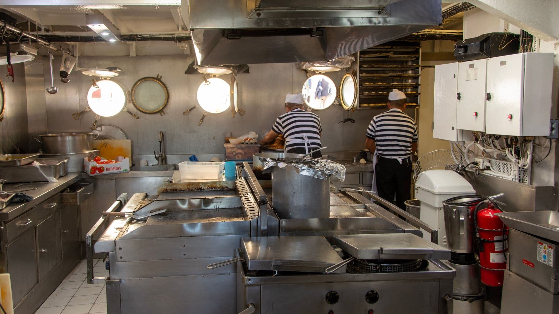 Two chefs in a ship's kitchen preparing meals with industrial equipment.