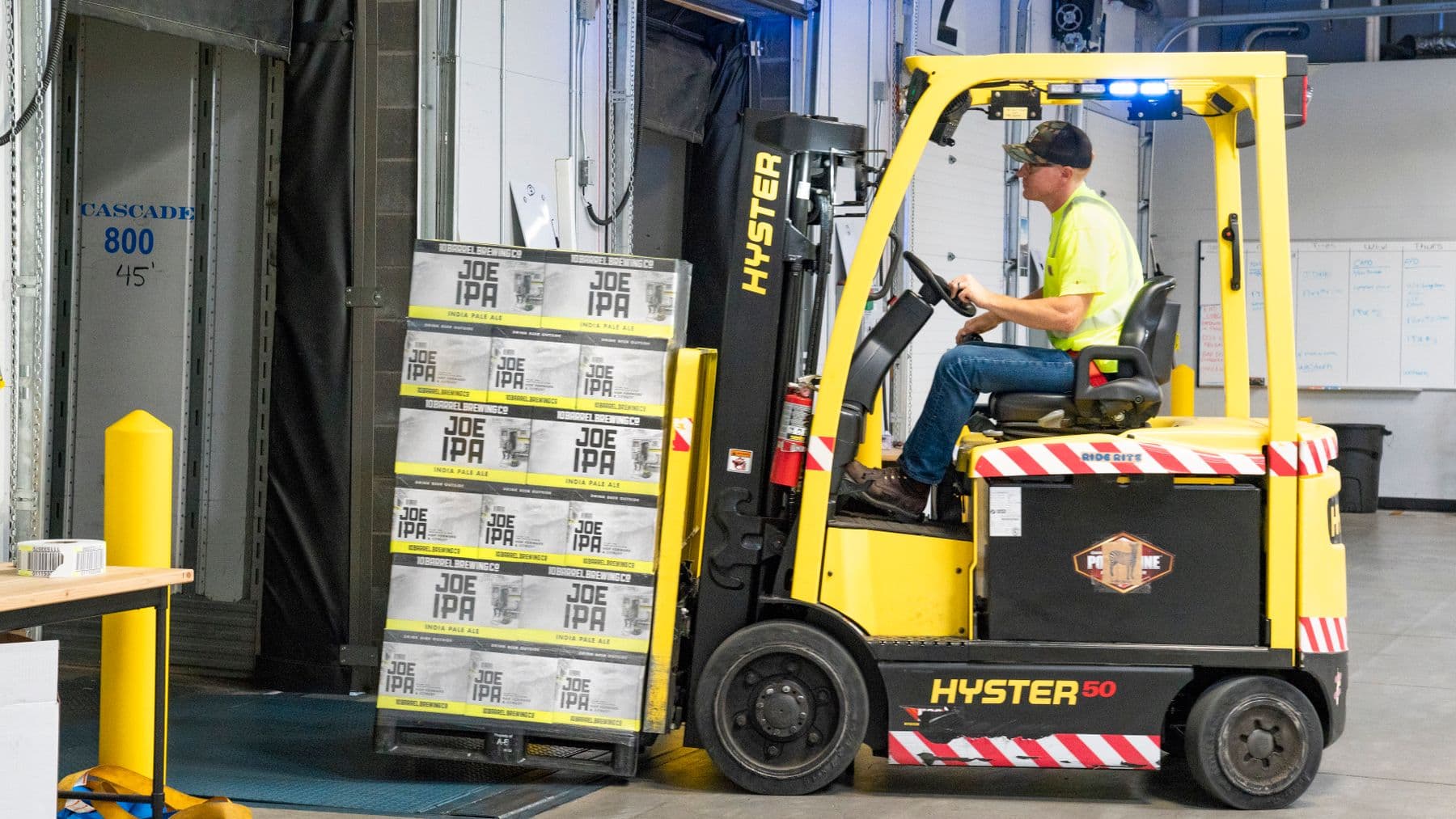 A worker drives a Hyster forklift moving Joe IPA boxes in a warehouse.